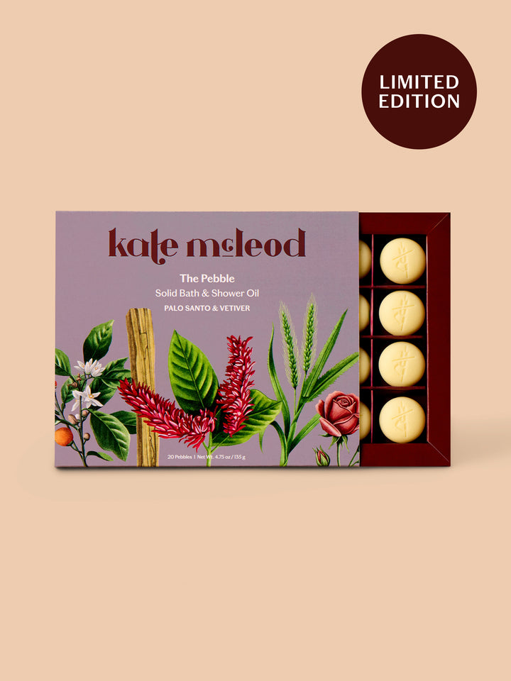 Kate McLeod 'The Pebble' solid bath and shower oil in a limited edition box, featuring botanical illustrations on a lavender background with pale yellow round bars visible in the tray, set against a warm beige background with a 'LIMITED EDITION' badge.