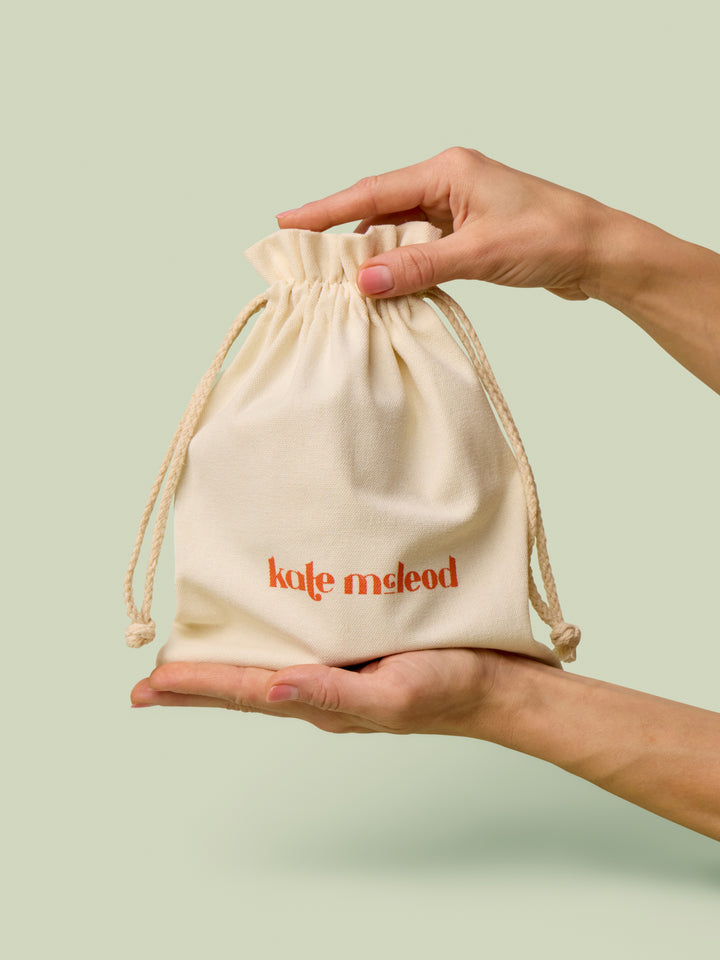 Hands holding a beige drawstring bag with Kate McLeod logo, the Gift Bag, on a light green background