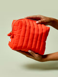 Person holding the Travel Bag, a poppy-orange quilted, zippered pouch, against a light green background.