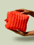 Person holding the Travel Bag, a poppy-orange quilted, zippered pouch, against a light green background.