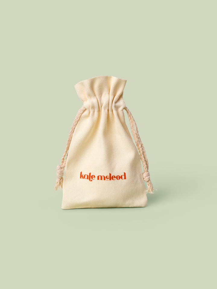 A beige drawstring bag with Kate McLeod logo, the Gift Bag, on a light green background