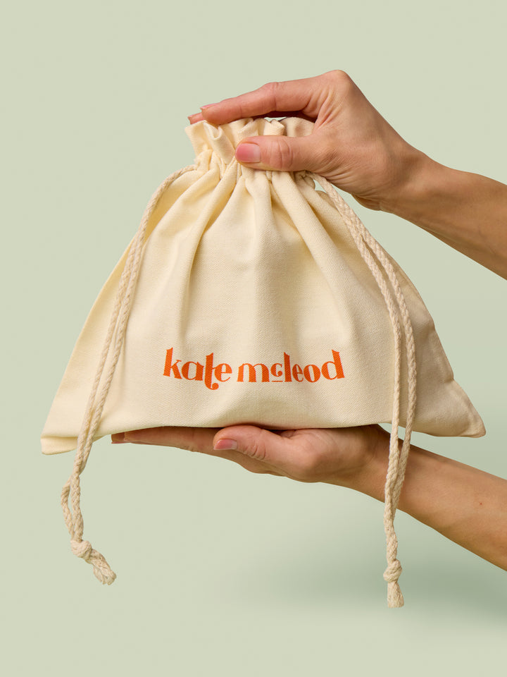 Large beige drawstring bag with Kate McLeod logo, the Large Gift Bag, held by hands against a light green background