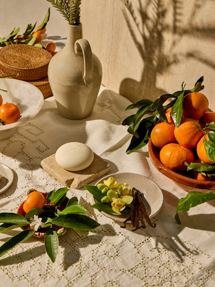 Kate McLeod Tangerine & Vanilla lotion bar on a stone plate with bowls full of oranges and vanilla flowers and vanilla bean stalks.