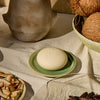 Kate McLeod Tonka Bean & Coconut lotion bar on a green plate with toasted coconut, tonka beans, and linen tablecloth.