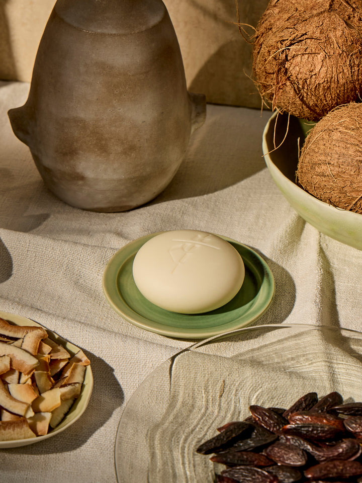 Kate McLeod Tonka Bean & Coconut lotion bar on a green plate with toasted coconut, tonka beans, and linen tablecloth.