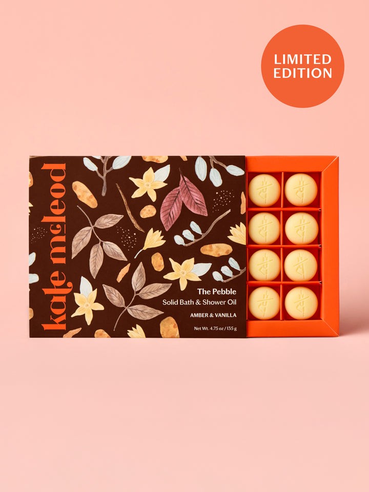 Box of Kate McLeod Amber & Vanilla solid bath and shower oil with brown floral illustrated packaging design on a light pink background.