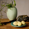 Amber & Vanilla Pebbles on a blue plate surrounded by vanilla and amber ingredients with a green vase and plant in the background.
