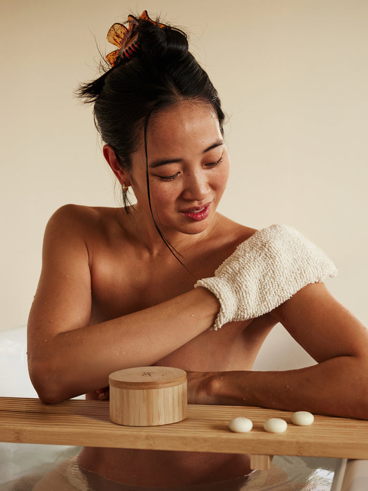 Woman using the body mitt to exfoliate her skin in the bath alongside the bamboo canister and sleep pebbles on a neutral background