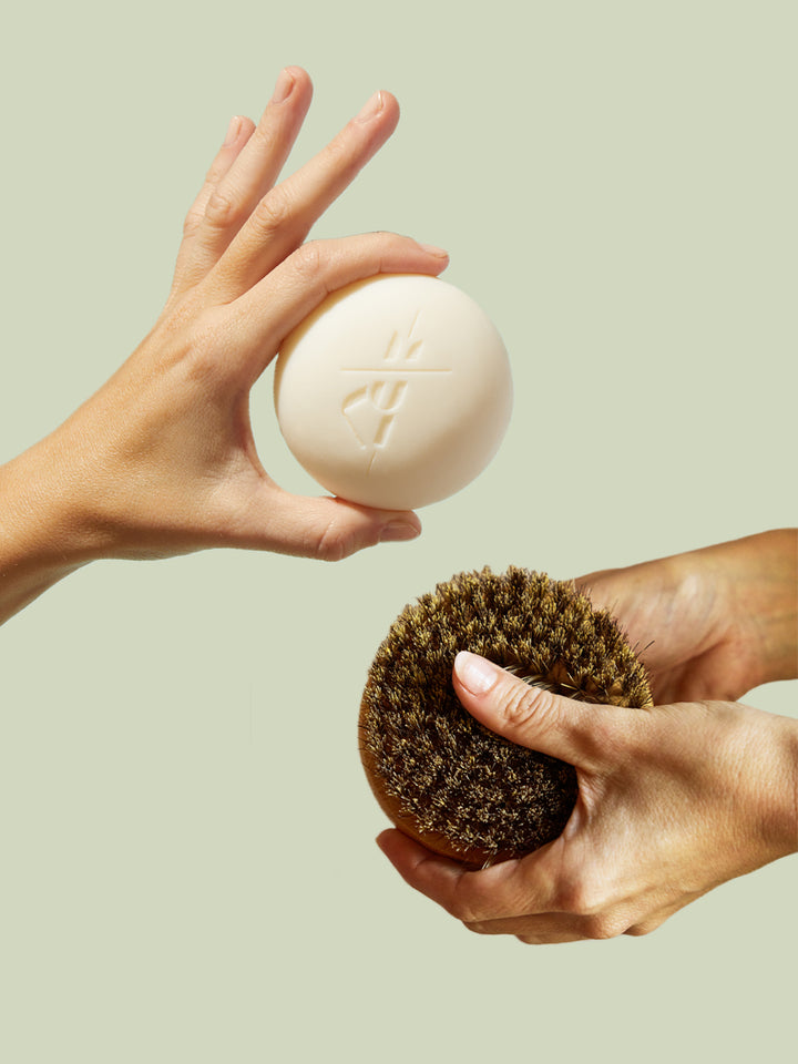 Two hands holding a bar of soap and a scrubber against a light green background