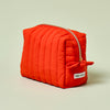 The Travel Bag, a poppy-orange quilted, zippered pouch, against a light green background.