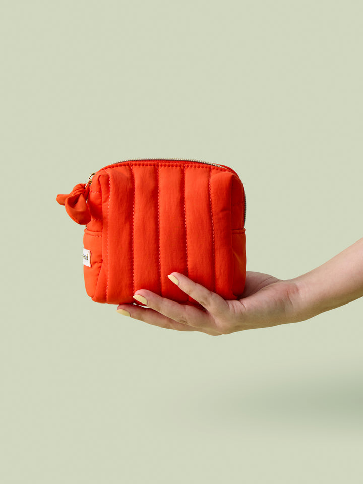 Hand holding Mini Travel Bag, a orange red poppy colored quilted zippered pouch, against a light green background.