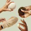 Hands holding various bath products including a loofah, scrubber, and bath bombs against a light green background.