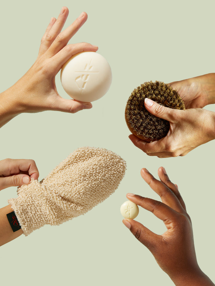 Hands holding various bath products including a loofah, scrubber, and bath bombs against a light green background.