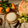 Kate McLeod Tangerine & Vanilla lotion bar with packaging on a wooden plate with ingredients and linens around