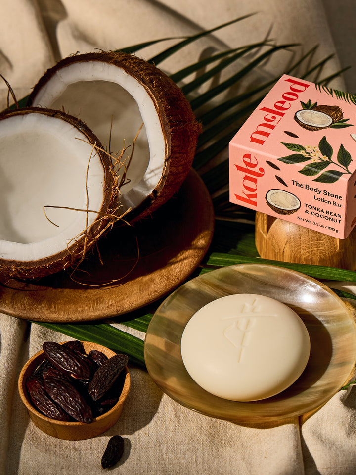 Kate McLeod Tonka Bean & Coconut lotion bar with packaging on a dish with ingredients and linens around
