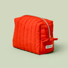 The Travel Bag, a poppy-orange quilted, zippered pouch, against a light green background.