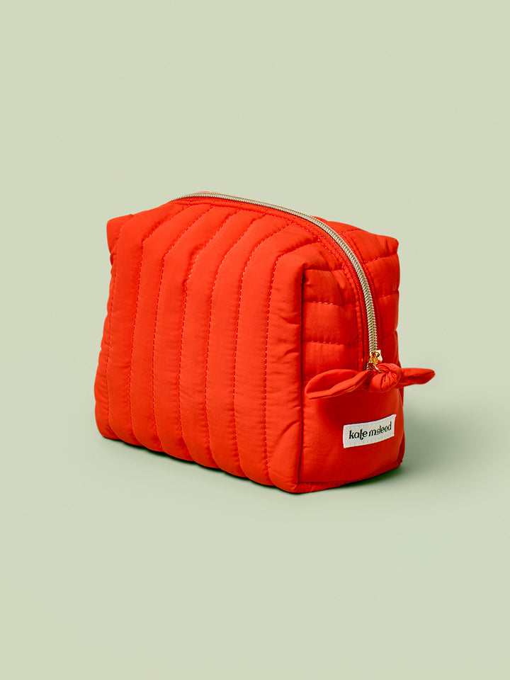 The Travel Bag, a poppy-orange quilted, zippered pouch, against a light green background.