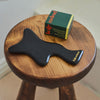 Black Body Gua Sha tool sitting atop a wooden stool next to the Recovery Stone on a neutral background.