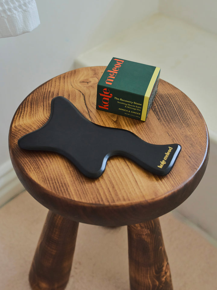 Black Body Gua Sha tool sitting atop a wooden stool next to the Recovery Stone on a neutral background.