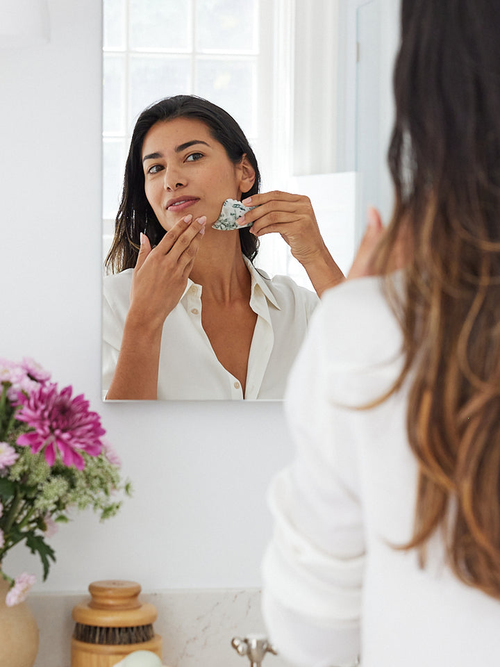 Woman the Gua Sha along her jawline in front of a mirror