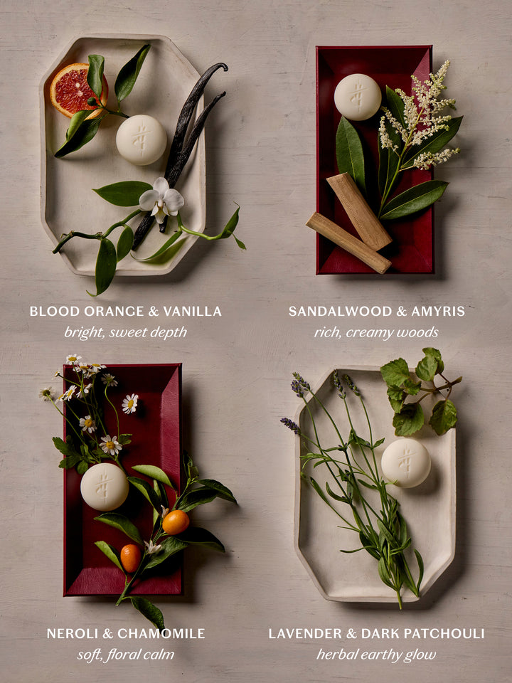 Four rectangular bath or body bars are arranged in a 2x2 grid on a light stone surface, each styled with botanical ingredients and text labels underneath. The products are: Blood Orange & Vanilla, Sandalwood & Amyris, Neroli & Chamomile, and Lavender & Dark Patchouli.