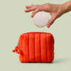 Hand holding a body stone above the poppy quilted mini travel bag on a light green background
