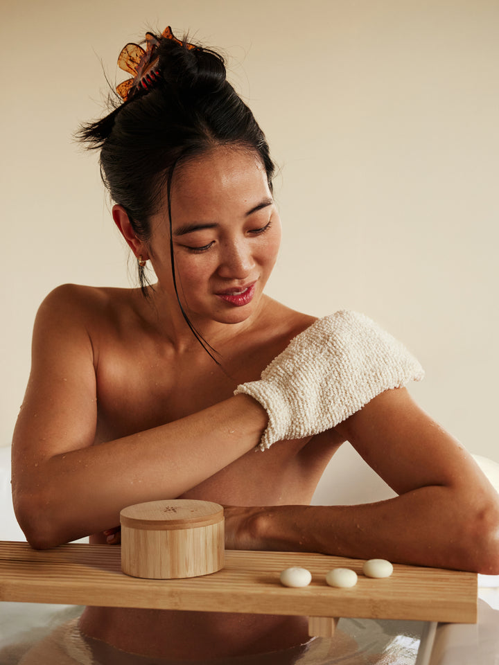 Woman exfoliating arms with the body mitt, sitting on a wooden bench.