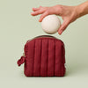 Hand placing a Mini Travel Case into a dark red quilted Mini Travel Bag on a light green background