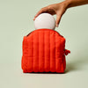 Hand placing the Full-Size Travel Case in ivory color into the Travel Bag, a poppy-orange quilted zippered pouch, on a light green background.
