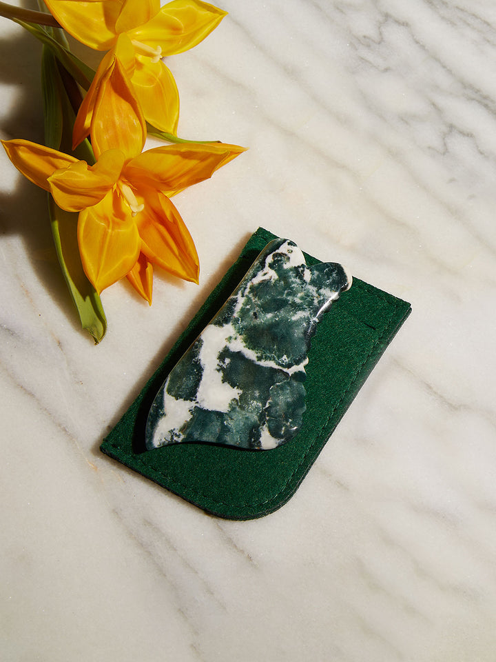 Face Gua Sha and green felt case on marble counter with yellow flowers.