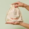 Hands holding a beige drawstring bag with Kate McLeod logo, the Gift Bag, on a light green background