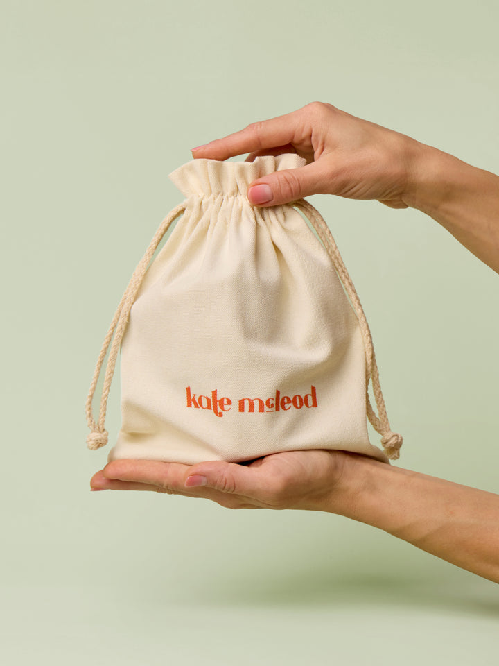 Hands holding a beige drawstring bag with Kate McLeod logo, the Gift Bag, on a light green background