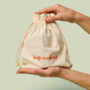 Hands holding a beige drawstring bag with Kate McLeod logo, the Gift Bag, on a light green background