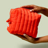 Person holding the Travel Bag, a poppy-orange quilted, zippered pouch, against a light green background.