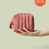 Hand holding Mini Travel Bag, a limited edition blush colored quilted zippered pouch, against a light green background.