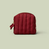 The Mini Travel Bag, a wine-red quilted zippered pouch, against a light green background.