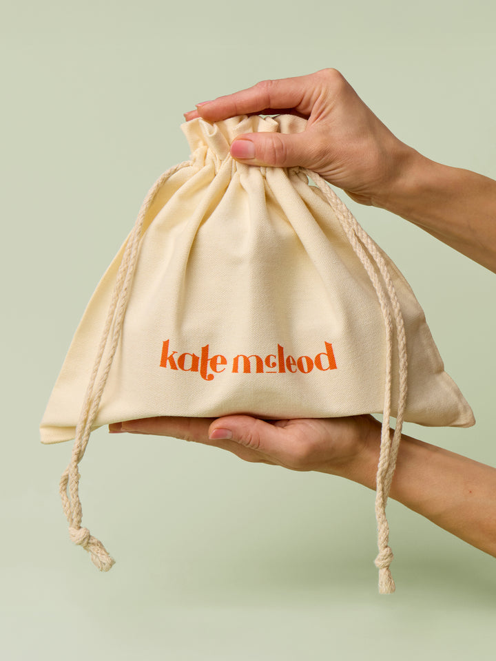 Large beige drawstring bag with Kate McLeod logo, the Large Gift Bag, held by hands against a light green background