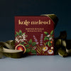 A boxed gift set from Kate McLeod titled 'Winter Rituals' with botanical illustrations on a dark red box, styled with olive green ribbon on a dark blue background.