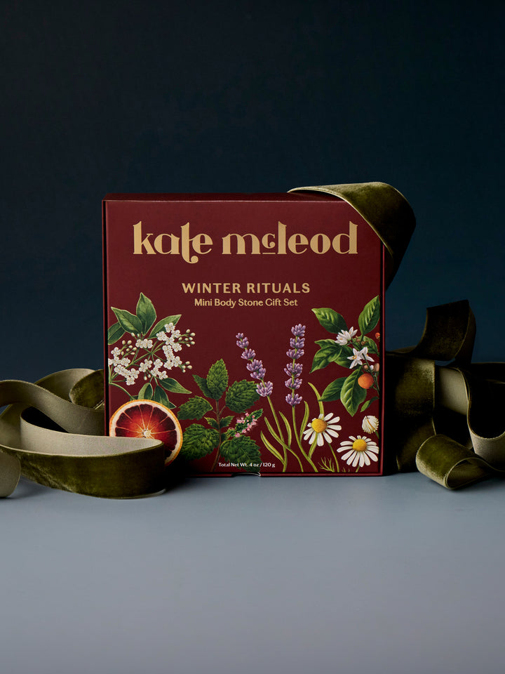 A boxed gift set from Kate McLeod titled 'Winter Rituals' with botanical illustrations on a dark red box, styled with olive green ribbon on a dark blue background.