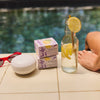 Stacked Kate McLeod Lavender Lemonade Body Stones styled next to the Travel Case. A red pair of sunglasses and refreshing lemonade sit nearby poolside.