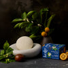 Lifestyle image of Jasmine & Sweet Orange Body Stone on a stone dish with packaging, oranges, and jasmine in a vase.