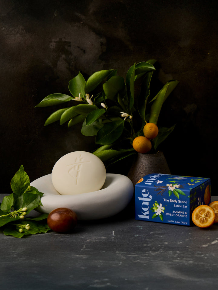 Lifestyle image of Jasmine & Sweet Orange Body Stone on a stone dish with packaging, oranges, and jasmine in a vase.