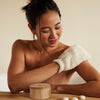 Woman using the body mitt to exfoliate her skin in the bath alongside the bamboo canister and sleep pebbles on a neutral background