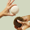 Two hands holding a bar of soap and a scrubber against a light green background
