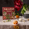 A styled tabletop scene featuring a boxed set labeled 'kale meleod winter rituals mini body ritual gift set' beside a large red rose in a glass vase, with citrus peels, a white stone, and small flowers on a linen surface against a dark textured backdrop.