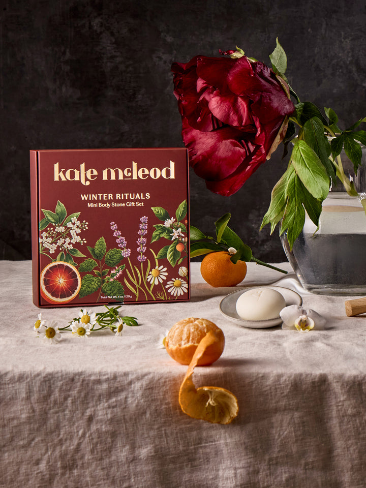 A styled tabletop scene featuring a boxed set labeled 'kale meleod winter rituals mini body ritual gift set' beside a large red rose in a glass vase, with citrus peels, a white stone, and small flowers on a linen surface against a dark textured backdrop.