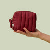 Hand holding Mini Travel Bag, a wine-red quilted zippered pouch, against a light green background.