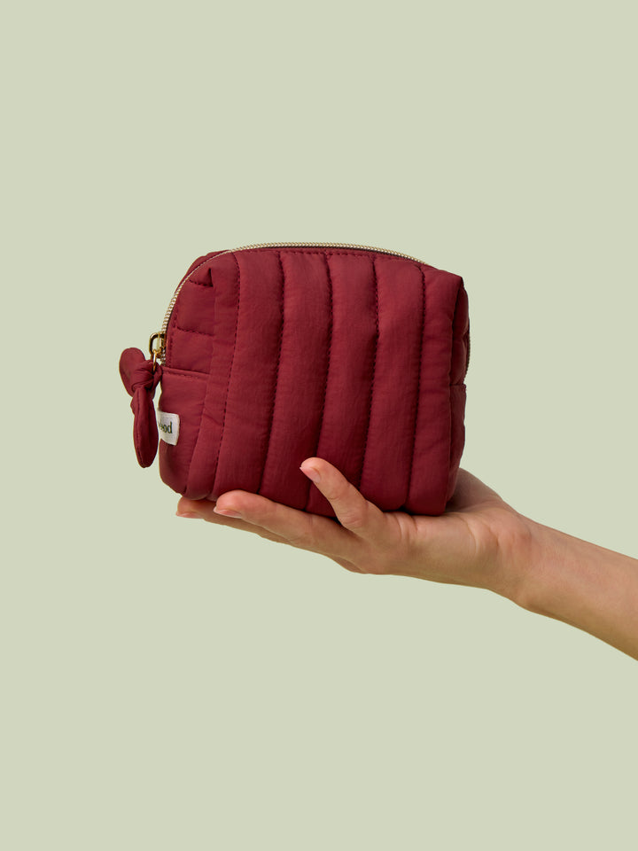 Hand holding Mini Travel Bag, a wine-red quilted zippered pouch, against a light green background.