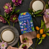 Kate McLeod Spring Awakening Body Stone Duo royal blue packaging with 2 white Body Stones on dishes, purple flowers, oranges, and a pink ribbon.