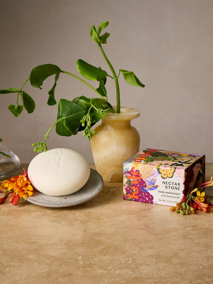 Nectar Stone on a plate with honeysuckle, plant in stone vase, and packaging on a beige table.